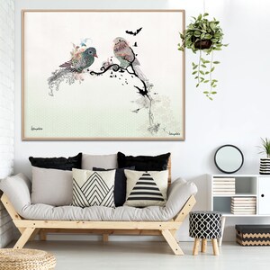 A framed art print featuring two stylized birds perched on a branch with a green and white background. The birds are decorated with intricate patterns and details. The print is framed in a light wood frame.