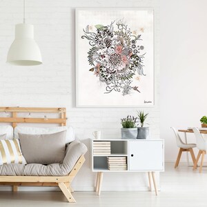 A white framed artwork with a black and white floral design with hints of pink and green. The artwork is hanging on a white brick wall above a light wood futon with two pillows, one with white and gold stripes. A white cabinet with a door and shelves is to the right of the futon. A white dining table with four white chairs is in the background.