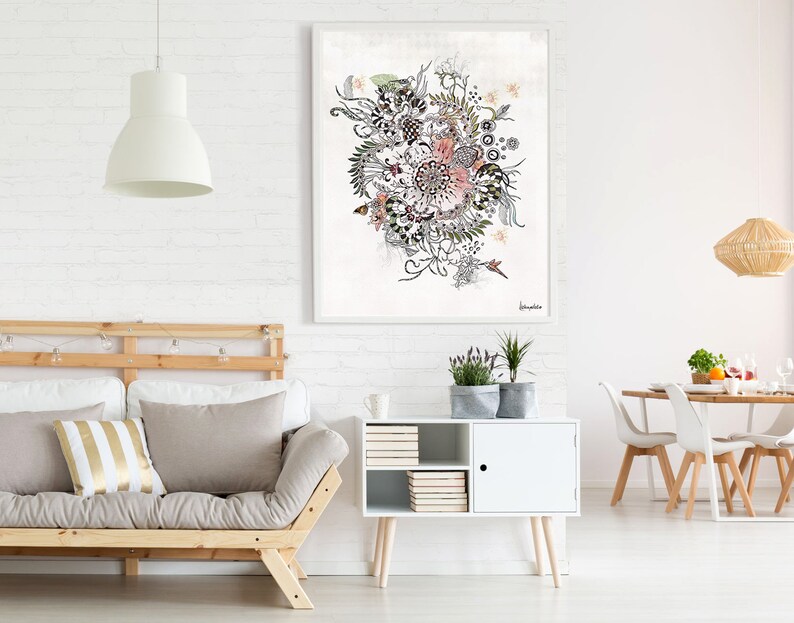 A white framed artwork with a black and white floral design with hints of pink and green. The artwork is hanging on a white brick wall above a light wood futon with two pillows, one with white and gold stripes. A white cabinet with a door and shelves is to the right of the futon. A white dining table with four white chairs is in the background.
