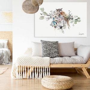 A watercolor painting of an owl perched on a branch with green leaves and flowers. The painting is framed and hanging on a white wall above a light gray couch with white throw pillows and a white blanket.