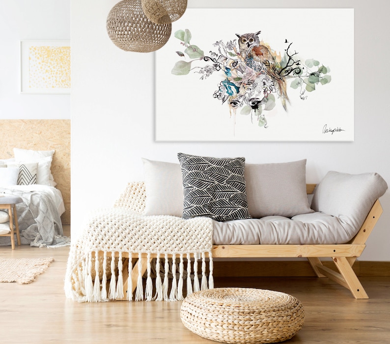 A watercolor painting of an owl perched on a branch with green leaves and flowers. The painting is framed and hanging on a white wall above a light gray couch with white throw pillows and a white blanket.