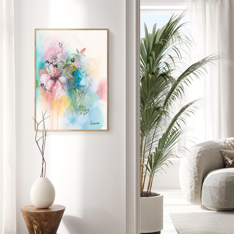 A framed watercolor painting of a pink flower with green leaves and blue and yellow watercolor washes in the background. The painting is hanging on a white wall in a room with a potted plant and a white couch.