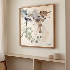 A framed watercolor painting features birds and eucalyptus branches. The artwork is in soft, muted tones of beige, brown, and green.  The birds are perched on branches, and the eucalyptus leaves are depicted with delicate brushstrokes. The painting has a light, airy feel, with a soft, watercolor wash background. The artist's signature is visible in the bottom right corner. The painting is displayed on a light beige wall, above a light wood shelf with two small beige cups.