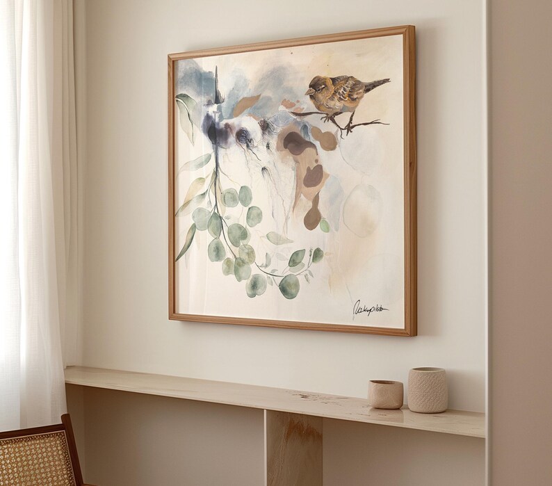 A framed watercolor painting features birds and eucalyptus branches. The artwork is in soft, muted tones of beige, brown, and green.  The birds are perched on branches, and the eucalyptus leaves are depicted with delicate brushstrokes. The painting has a light, airy feel, with a soft, watercolor wash background. The artist's signature is visible in the bottom right corner. The painting is displayed on a light beige wall, above a light wood shelf with two small beige cups.