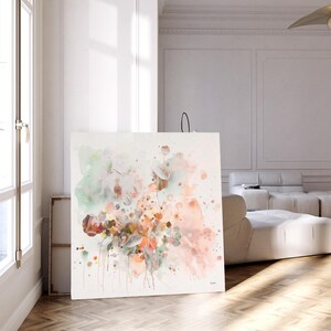 Large square canvas art featuring an abstract floral design. The artwork showcases a blend of soft colors, including peach, pink, green, and brown, with paint splatters. The piece is displayed in a bright, modern room with natural light.