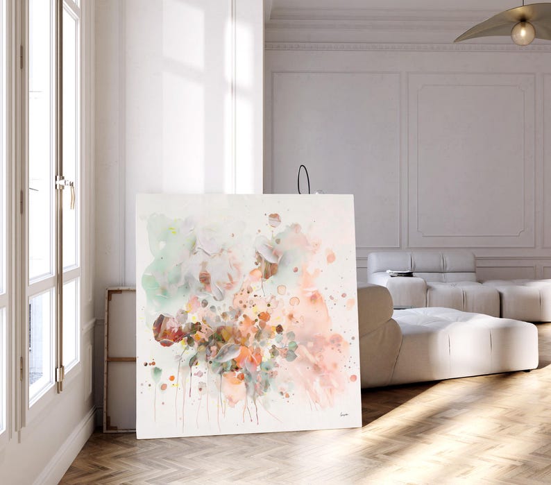 Large square canvas art featuring an abstract floral design. The artwork showcases a blend of soft colors, including peach, pink, green, and brown, with paint splatters. The piece is displayed in a bright, modern room with natural light.