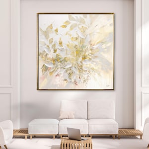 Large abstract painting with a gold frame. The artwork features a floral design in shades of white, cream, and gold. The painting is displayed above a white sofa and ottoman in a modern living room setting.
