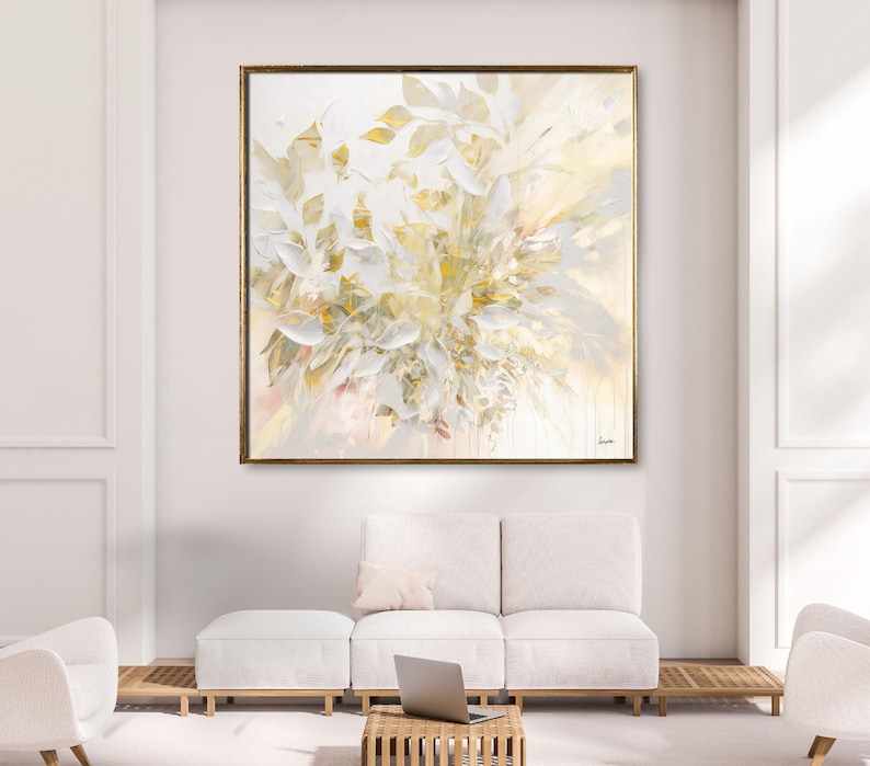 Large abstract painting with a gold frame. The artwork features a floral design in shades of white, cream, and gold. The painting is displayed above a white sofa and ottoman in a modern living room setting.