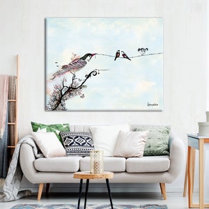 A watercolor painting of birds on a wire with a blue sky background. The painting is framed and hanging on a white wall above a white couch with green and white pillows. The couch is in a living room with a wooden side table and a ladder with a blanket on it.
