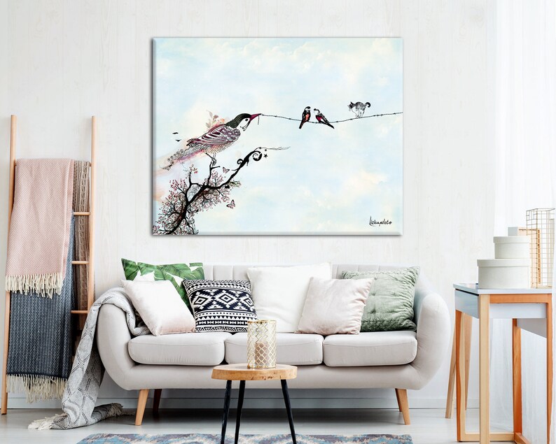 A watercolor painting of birds on a wire with a blue sky background. The painting is framed and hanging on a white wall above a white couch with green and white pillows. The couch is in a living room with a wooden side table and a ladder with a blanket on it.