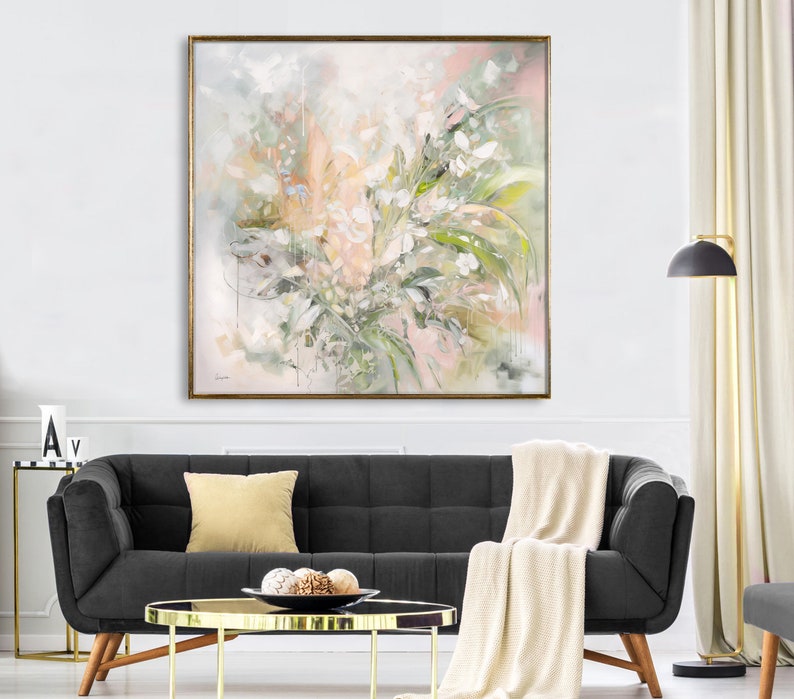 A large abstract painting with a gold frame, featuring a light pink, green, and white floral design. The painting is hanging on a white wall above a black sofa with a white throw blanket and a gold coffee table.