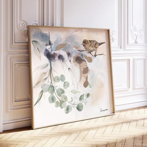 A large watercolor painting on canvas features a sparrow perched on a eucalyptus branch. The artwork is in muted earth tones of beige, brown, and green, with soft watercolor washes creating a dreamy atmosphere. The bird is realistically depicted, while the leaves and background are more abstract. The painting is framed in a light gold frame and is displayed against a white wall. The artist's signature is visible in the bottom right corner.