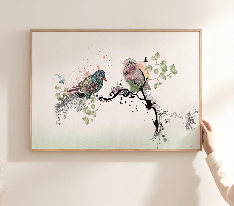 A framed wall art print featuring two stylized birds perched on a black ink-style branch. The birds are decorated with intricate patterns in shades of blue, green, and pink. Soft watercolor splashes and green leaves surround the birds.