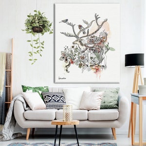 A watercolor-style canvas print featuring a deer surrounded by birds and floral elements. The artwork is in shades of beige, gray, and green, with hints of pink and orange. The deer is centrally positioned, with its antlers extending outward. The birds are perched on the antlers and within the foliage. The overall style is whimsical and nature-inspired. The artwork is displayed on a wall above a light beige sofa with green and patterned throw pillows.