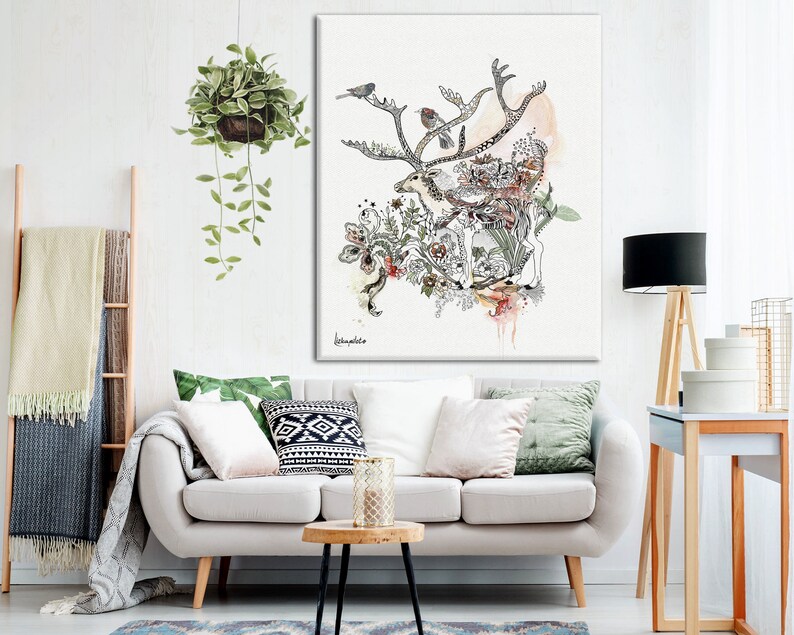 A watercolor-style canvas print featuring a deer surrounded by birds and floral elements. The artwork is in shades of beige, gray, and green, with hints of pink and orange. The deer is centrally positioned, with its antlers extending outward. The birds are perched on the antlers and within the foliage. The overall style is whimsical and nature-inspired. The artwork is displayed on a wall above a light beige sofa with green and patterned throw pillows.