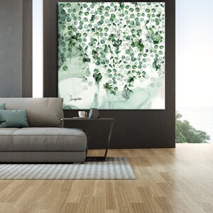 A large, framed painting of green leaves and vines hanging on a wall. The painting is in a modern style and has a white background. The leaves are a variety of shades of green and are arranged in a cascading pattern. The painting is hanging above a gray couch and a black coffee table. The couch is in a living room with a large window overlooking a body of water.