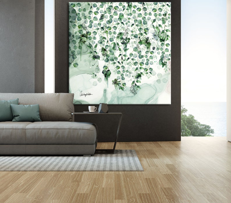 A large, framed painting of green leaves and vines hanging on a wall. The painting is in a modern style and has a white background. The leaves are a variety of shades of green and are arranged in a cascading pattern. The painting is hanging above a gray couch and a black coffee table. The couch is in a living room with a large window overlooking a body of water.