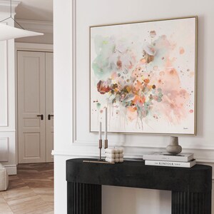 Abstract floral watercolor painting in a gold frame. The artwork features soft pastel colors, including pink, green, and orange, with a white background. The painting is displayed above a black console table with decorative items.