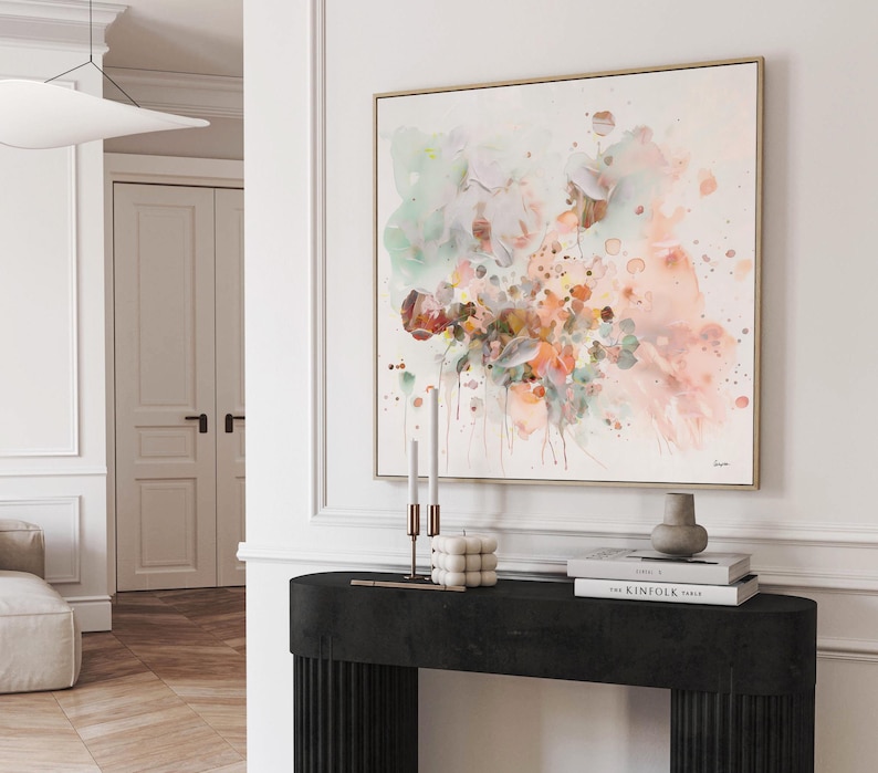 Abstract floral watercolor painting in a gold frame. The artwork features soft pastel colors, including pink, green, and orange, with a white background. The painting is displayed above a black console table with decorative items.