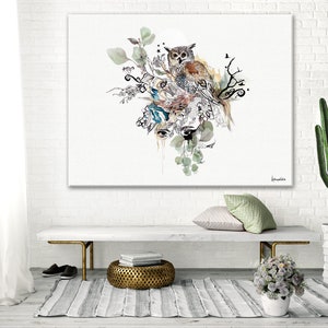 A watercolor painting of an owl perched on a branch with green leaves and flowers. The owl is brown and white with black eyes. The painting is on a white canvas with a white background. The painting is hanging on a white brick wall above a white bench with a gold metal base. There is a small potted plant with white flowers on the bench. There is a cactus plant in a white pot on the right side of the image. The painting is signed 'devandelle' in the bottom right corner.
