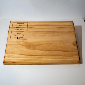 May include: A rectangular wooden cutting board with a light brown finish. The board features the quote "There is no sincerer love than the love of food" by George Bernard Shaw, etched in black. The wood grain is visible.