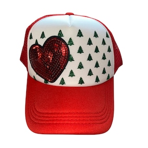 May include: Red trucker hat with a white front panel featuring a sequined heart and green Christmas trees. The hat has a red brim and a red mesh back, perfect for a festive look.