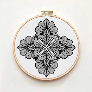 Peut inclure: Broderie au point de croix terminée, présentée dans un cercle en bois. Le motif floral symétrique est réalisé en nuances de gris et de noir. L'ouvrage est centré sur un fond de tissu blanc, créant un contraste saisissant.