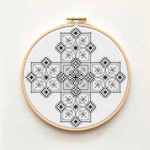 Peut inclure: Une broderie au point de croix en noir et blanc, représentant un motif géométrique, présentée dans un cercle à broder en bois. Le motif est sur un tissu blanc, avec des éléments symétriques et détaillés.