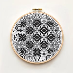 May include: A black and white cross-stitch embroidery in a wooden hoop. The design features a geometric pattern with floral-like shapes. The embroidery is stretched taut within the round frame, showcasing the intricate details of the needlework.