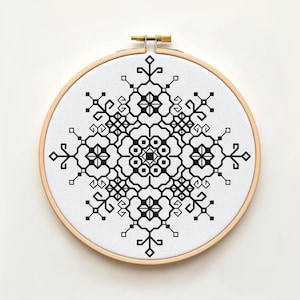 Peut inclure: Un tambour à broder circulaire en bois contenant un tissu blanc avec un motif au point de croix noir. Le design présente un motif géométrique symétrique en forme de flocon, composé de lignes, de carrés et de losanges entrelacés.