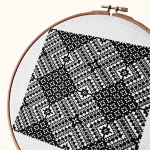 Peut inclure: Un tambour à broder circulaire en bois contenant un tissu Aida blanc avec un motif de point de croix noir. Le design présente un motif géométrique répétitif de carrés et de losanges, créant une grille symétrique, complexe et contrastée.