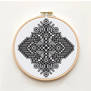 May include: A finished cross-stitch piece with a geometric black and white pattern. The design is set on white fabric, held within a wooden embroidery hoop. The pattern includes squares, rectangles, and floral-like shapes.