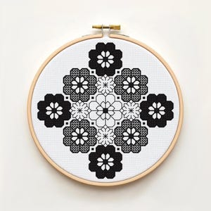Peut inclure: Un tambour à broder circulaire en bois contenant un tissu blanc avec un motif de point de croix noir. Le design présente une disposition symétrique de formes florales noires pleines et détourées, créant un motif géométrique en étoile.