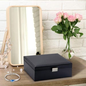May include: A rectangular black jewelry box with a silver-toned metal clasp, positioned next to a silver bracelet with a blue stone, a light-colored wooden mirror, and a clear glass vase containing a bouquet of pink roses.