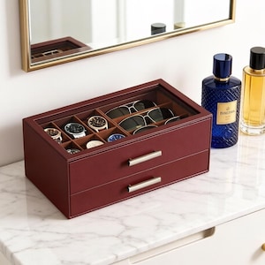 May include: A burgundy leather watch and sunglasses organizer with a glass top and two drawers. The top section holds watches and sunglasses. Two perfume bottles sit to the right of the organizer. A mirror is in the background.