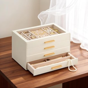 May include: A cream-colored jewelry box with a glass top lid, three drawers, and gold-toned horizontal handles. The interior is lined with beige fabric, featuring compartments for rings, watches, and necklaces, with one drawer pulled open.