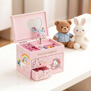 May include: Pink wooden jewelry box with a heart-shaped mirror and a unicorn figurine inside. The box has drawers and is decorated with unicorn and rainbow illustrations. The text "You are magical" is printed on the front. A teddy bear and a white bunny plush toy are next to the box.