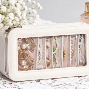 May include: A cream-colored rectangular jewelry case with a clear window displaying various necklaces and earrings. The case is zippered and sits on a white lace surface, with two perfume bottles in the background.
