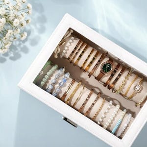May include: A white jewelry box filled with various bracelets and a watch. The bracelets are gold, silver, and beaded, with some featuring engraved text. The watch has a brown leather strap and a green face. The box is on a light blue surface.