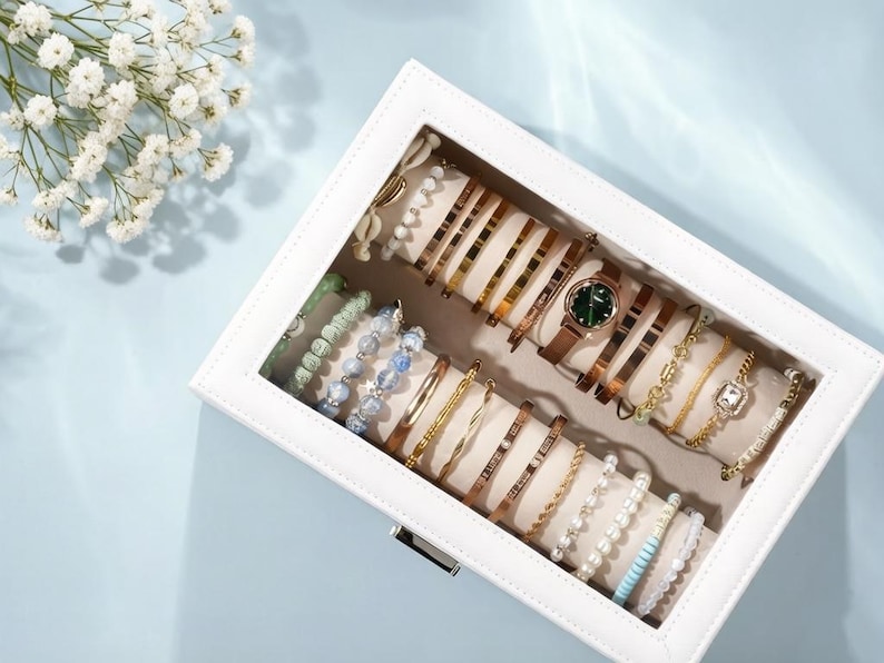 May include: A white jewelry box filled with various bracelets and a watch. The bracelets are gold, silver, and beaded, with some featuring engraved text. The watch has a brown leather strap and a green face. The box is on a light blue surface.
