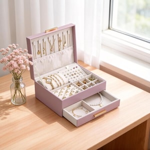 May include: A lavender jewelry box with multiple compartments and a pull-out drawer. The box is open, revealing necklaces, bracelets, rings, and earrings. Gold-colored hardware accents the box. A vase of pink flowers sits nearby.