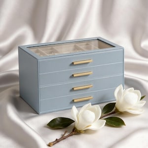 May include: A light blue jewelry box with a clear glass top lid and three horizontal drawers, each featuring a slim gold-toned metal handle. The rectangular storage unit is positioned next to two white magnolia flowers with green leaves.