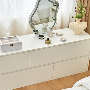 May include: A white dresser with a wavy-edged mirror, a decorative vase with flowers, and various cosmetic products. A jewelry box sits on the dresser, reflecting a clean and organized aesthetic. The room is well-lit, suggesting a bright and airy space.