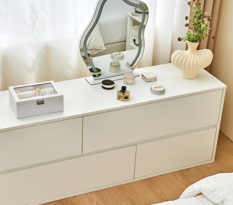 May include: A white dresser with a wavy-edged mirror, a decorative vase with flowers, and various cosmetic products. A jewelry box sits on the dresser, reflecting a clean and organized aesthetic. The room is well-lit, suggesting a bright and airy space.
