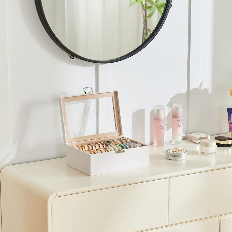 May include: A white jewelry box with a clear glass top, filled with various rings and bracelets. The box is open, revealing the contents. The jewelry box is on a white dresser, with a mirror in the background.