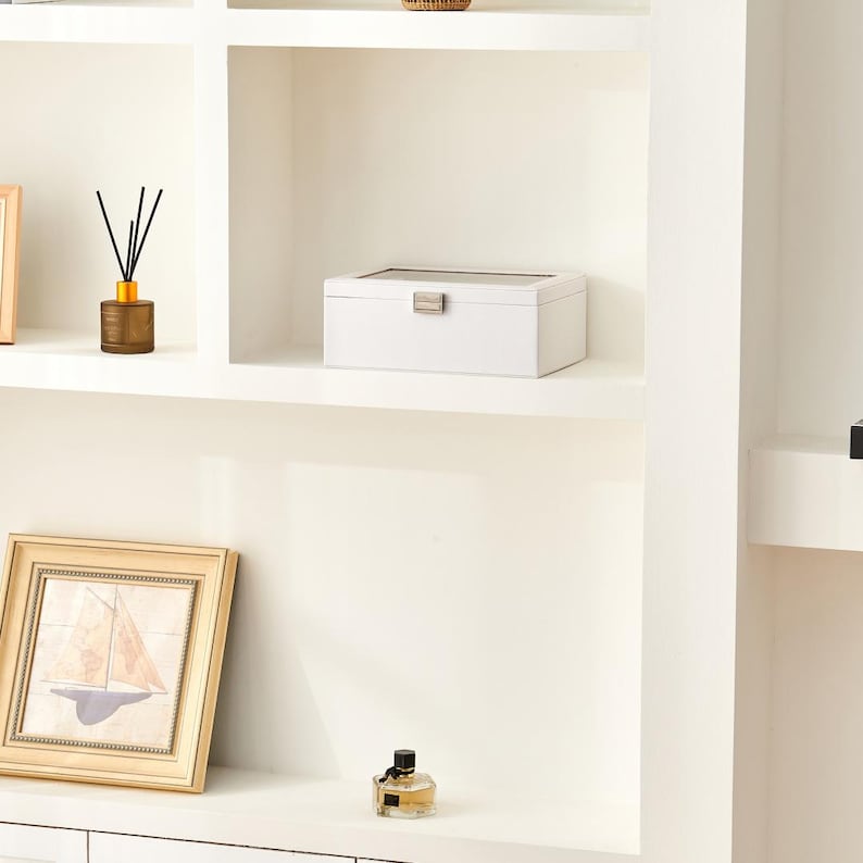May include: A white jewelry box with a rectangular shape and a silver clasp sits on a white shelf. A framed picture of a sailboat, a brown diffuser, and a perfume bottle are also on the shelf.