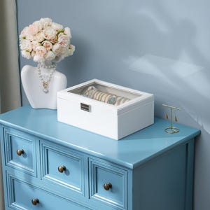 May include: A white jewelry box with a clear top sits on a light blue dresser. The box contains several bracelets and watches. A white bust with a floral arrangement and a necklace is to the left.