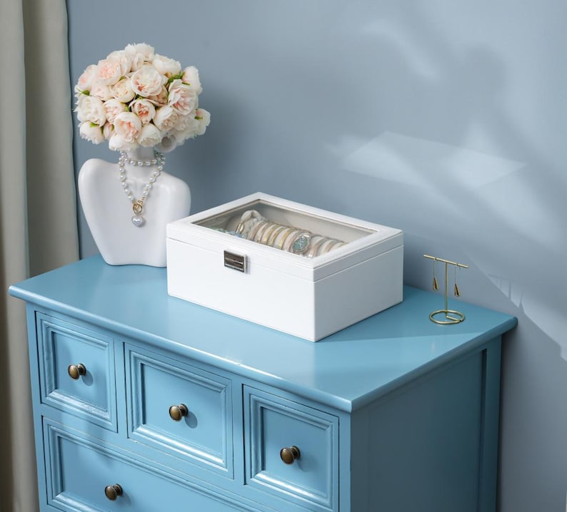 May include: A white jewelry box with a clear top sits on a light blue dresser. The box contains several bracelets and watches. A white bust with a floral arrangement and a necklace is to the left.