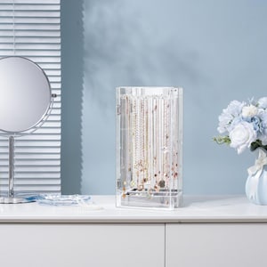 May include: A clear acrylic jewelry organizer filled with necklaces and other jewelry. A round, silver-framed mirror and a light blue vase with a bouquet of flowers are also on the white surface. The background is a light blue wall and a window with white blinds.