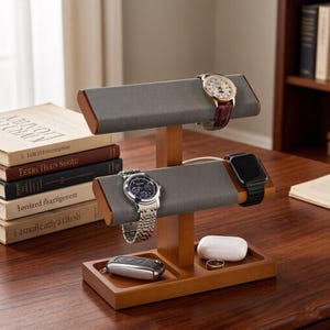May include: A two-tier wooden watch stand with grey fabric-covered bars for displaying timepieces. The base features two recessed compartments holding a silver key fob, a white charging case, and a metal ring. Three watches are displayed.